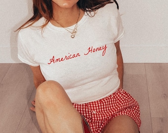 May include: A woman wearing a white t-shirt with the text "American Honey" in red script. She is wearing red and white gingham shorts and red cowboy boots with white stars.