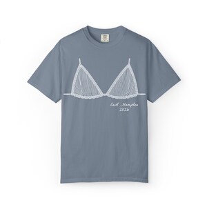 May include: A dusty blue t-shirt featuring a white line drawing of a bikini top. The bikini top design has vertical stripes and a scalloped edge. The words "East Hampton 2026" are written below the bikini top in a script font.