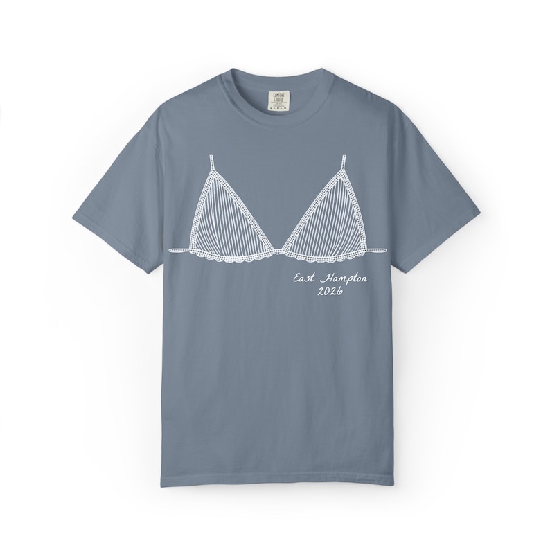 May include: A dusty blue t-shirt featuring a white line drawing of a bikini top. The bikini top design has vertical stripes and a scalloped edge. The words "East Hampton 2026" are written below the bikini top in a script font.