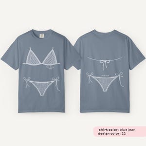 May include: Blue jean colored t-shirts with white line drawings of a bikini top and bottom on the front and back. The front of the shirt has the text "Palm Springs 2023". The back of the shirt has the text "Bikini".