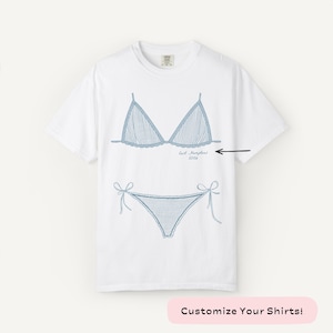 May include: White t-shirt featuring a blue line drawing of a bikini top and bottom. The bikini design includes vertical stripes and tie details. The text "East Hampton 2026" is below the bikini top. The bottom of the image has the text "Customize Your Shirts!"