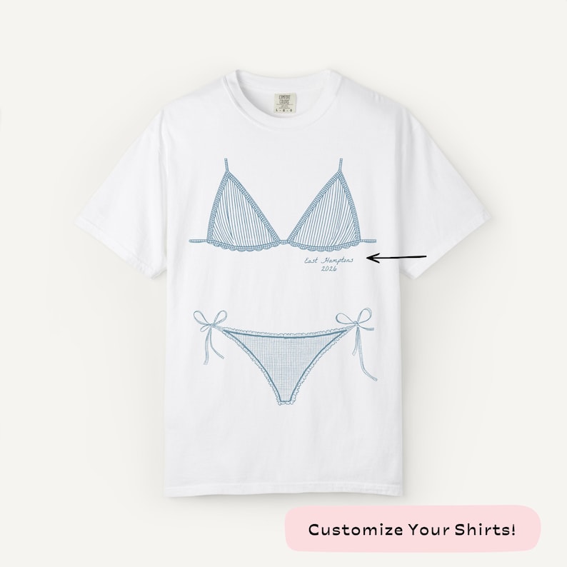 May include: White t-shirt featuring a blue line drawing of a bikini top and bottom. The bikini design includes vertical stripes and tie details. The text "East Hampton 2026" is below the bikini top. The bottom of the image has the text "Customize Your Shirts!"