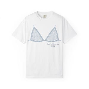 May include: White t-shirt featuring a blue line drawing of a bra. The bra design has vertical stripes and a scalloped edge. The text "East Hampton 2026" is written in cursive below the bra.
