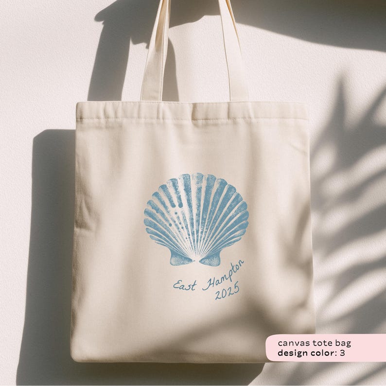 May include: Cream-colored canvas tote bag featuring a large blue seashell design. The words "East Hampton 2025" are printed below the seashell. The bag has two cream-colored handles.