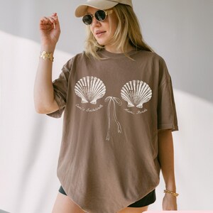 May include: Espresso-colored t-shirt featuring a seashell design with the text "Cami's Bachelorette, Miami, Florida" in white. The design includes two large seashells and a bow. The shirt is a relaxed fit.