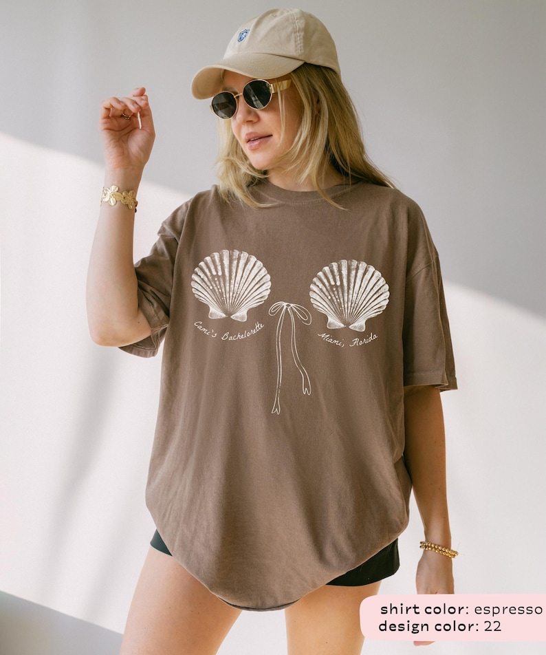 May include: Espresso-colored t-shirt featuring a seashell design with the text "Cami's Bachelorette, Miami, Florida" in white. The design includes two large seashells and a bow. The shirt is a relaxed fit.
