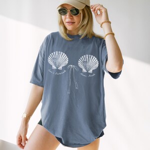 May include: Blue jean colored t-shirt with a white seashell design. The design includes the text "Cari's Bachelorette Miami, Florida". The shirt has short sleeves and a relaxed fit. The model is wearing a beige cap and sunglasses.