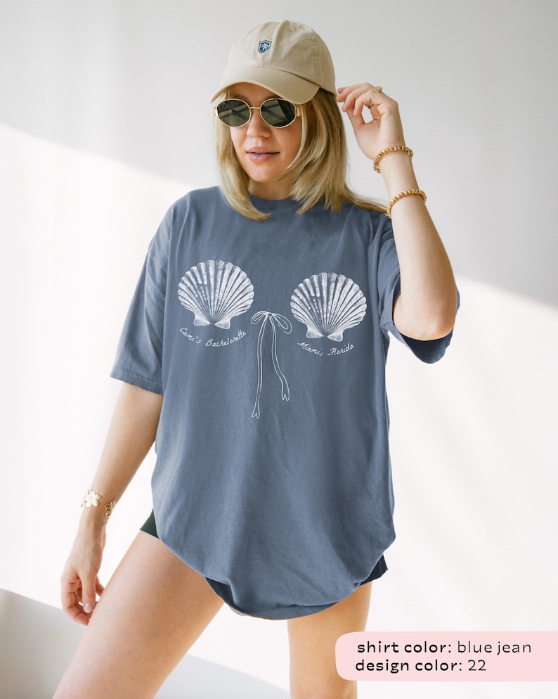 May include: Blue jean colored t-shirt with a white seashell design. The design includes the text "Cari's Bachelorette Miami, Florida". The shirt has short sleeves and a relaxed fit. The model is wearing a beige cap and sunglasses.