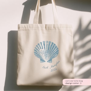May include: Cream-colored canvas tote bag featuring a light blue seashell design. The words "East Hampton 2026" are written below the seashell. The bag has two cream-colored handles.