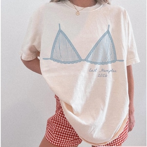 May include: Ivory-colored t-shirt featuring a blue striped bra design. The text "East Hampton 2026" is printed below the bra graphic. The model is wearing red and white checkered shorts. The shirt is a casual, relaxed fit.