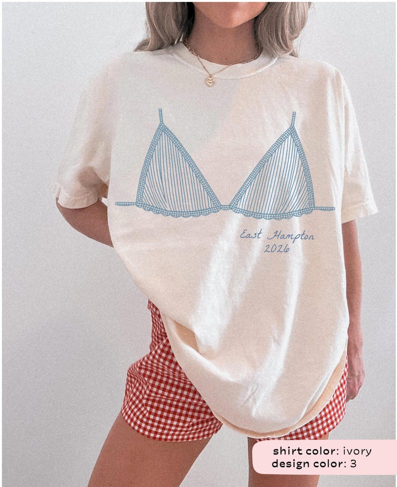 May include: Ivory-colored t-shirt featuring a blue striped bra design. The text "East Hampton 2026" is printed below the bra graphic. The model is wearing red and white checkered shorts. The shirt is a casual, relaxed fit.
