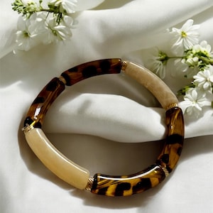 Può includere: Un braccialetto spesso con segmenti alternati in tartaruga e acrilico color crema. Il braccialetto è impreziosito da metallo color oro ed è esposto su uno sfondo bianco con fiori bianchi.