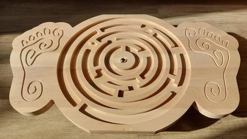 May include: A wooden maze game with a circular maze in the center and two foot-shaped pieces on either side. The maze has a small metal ball in the center.