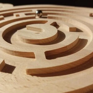 May include: A wooden maze with a metal ball in the center. The maze is made of concentric circles with a complex pattern of paths.