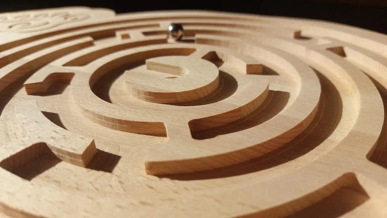 May include: A wooden maze with a metal ball in the center. The maze is made of concentric circles with a complex pattern of paths.