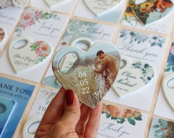 Puede incluir: Abrebotellas de cerámica blanco en forma de corazón con una foto de una pareja en un campo. La fecha 04 05 22 está impresa en el abrebotellas. El abrebotellas está rodeado de otros favores de boda con diseños florales y el texto "Gracias por celebrar con nosotros".
