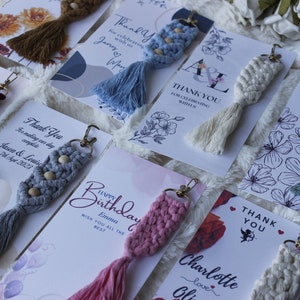 May include: Assortment of colorful macrame keychains with wooden beads and tassels. Each keychain is displayed on a card with a personalized message, such as "Thank You", "Happy Birthday", and "Thank You for celebrating with us".
