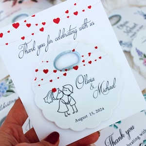 May include: White wedding favor tag with a red heart design and a bottle opener. The tag says "Thank you for celebrating with us" and has the couple's names, Olivia & Michael, and the wedding date, August 15, 2024.