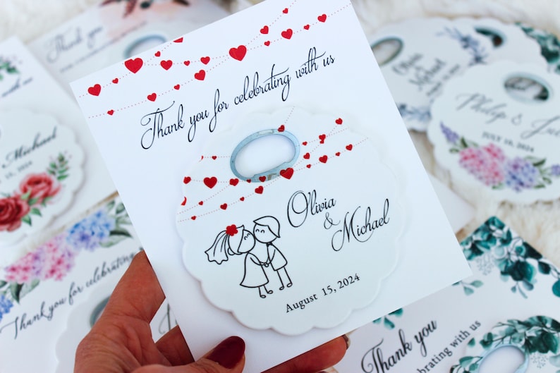 May include: White wedding favor tag with a red heart design and a bottle opener. The tag says "Thank you for celebrating with us" and has the couple's names, Olivia & Michael, and the wedding date, August 15, 2024.