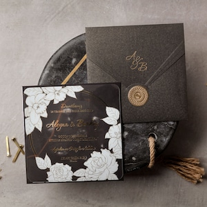May include: Elegant black wedding invitation suite with gold accents. Includes a square invitation with floral design, a matching envelope with a monogram, and a wax seal. Stationery is arranged on a marble-like surface with pencils and decorative elements.