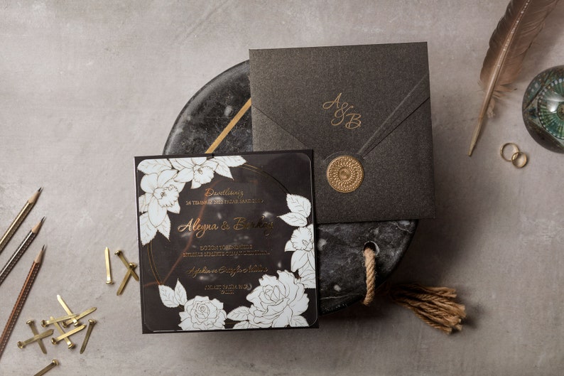 May include: Elegant black wedding invitation suite with gold accents. Includes a square invitation with floral design, a matching envelope with a monogram, and a wax seal. Stationery is arranged on a marble-like surface with pencils and decorative elements.