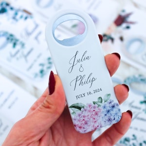 Pode incluir: Um abridor de garrafas azul claro com fundo branco e design floral. O abridor de garrafas tem os nomes "Julia & Philip" e a data "JULY 10, 2024" impressos nele.