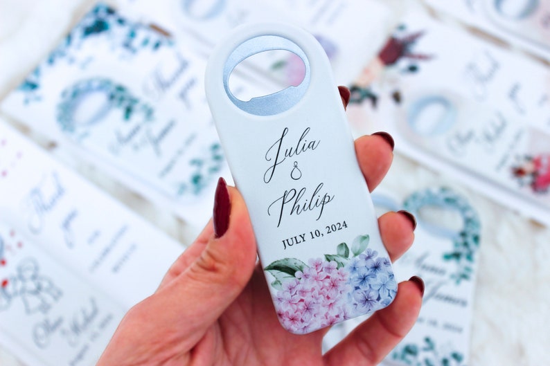 Pode incluir: Um abridor de garrafas azul claro com fundo branco e design floral. O abridor de garrafas tem os nomes "Julia & Philip" e a data "JULY 10, 2024" impressos nele.