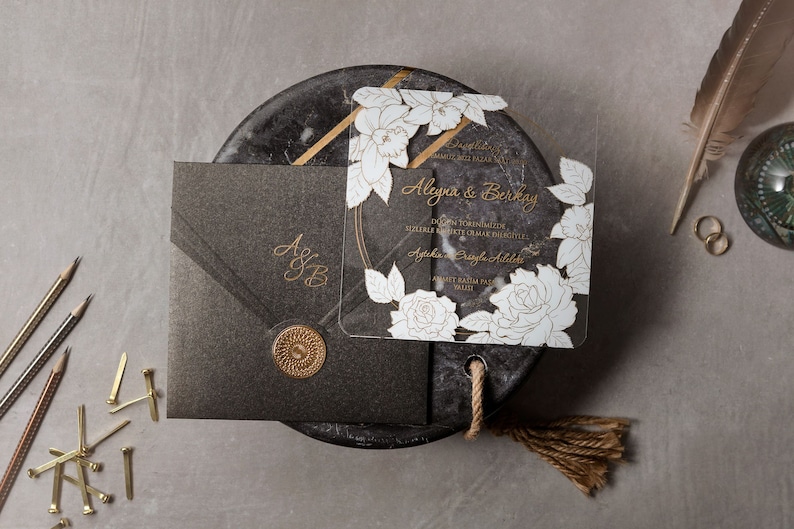 May include: Elegant wedding invitation suite featuring a dark gray envelope with gold initials and a floral-adorned transparent card. The invitation includes text in gold, a decorative seal, and a floral design. The suite is set on a textured surface.