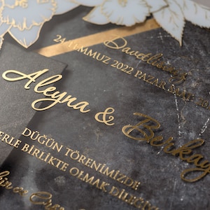 May include: Elegant wedding invitation with gold lettering on a dark gray background. The invitation includes the names "Aleyna & Berkay" and event details. Floral accents in white and gold add a decorative touch.