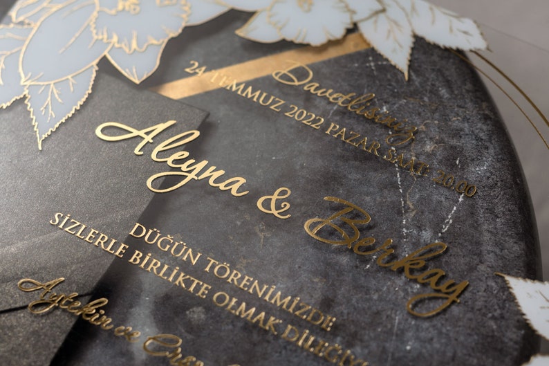 May include: Elegant wedding invitation with gold lettering on a dark gray background. The invitation includes the names "Aleyna & Berkay" and event details. Floral accents in white and gold add a decorative touch.