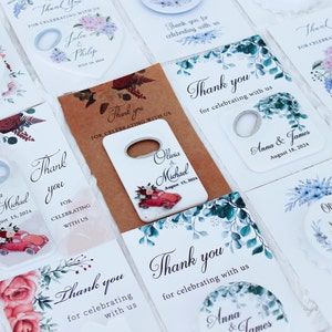 May include: Assortment of wedding favor tags with floral designs and bottle opener favors. The tags feature the names of the couples and their wedding dates. The bottle openers are white with a red car and floral designs.