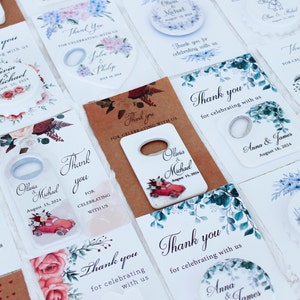 Pode incluir: Conjunto de abridores de garrafas para lembrancinhas de casamento com v&aacute;rios desenhos florais e os nomes dos casais. Os abridores de garrafas s&atilde;o de metal e t&ecirc;m um fundo branco com uma mensagem de agradecimento.