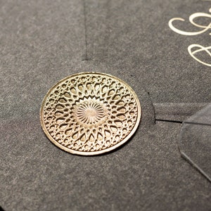 May include: A gold-colored, circular embellishment with an intricate, openwork design. The decorative element is placed on a dark gray surface, possibly a card or invitation. The initials "A & B" are visible in gold script.