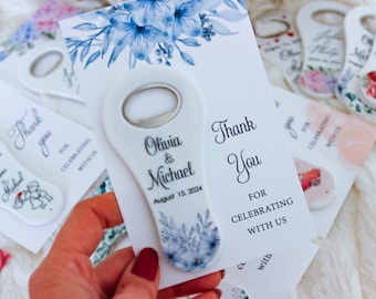 May include: White ceramic bottle opener with a blue floral design and the text "Olivia & Michael" and "August 15, 2024". The opener is attached to a white card with the text "Thank You for Celebrating with Us".