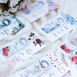 Pode incluir: Conjunto de seis abridores de garrafas de casamento com diferentes designs florais. Cada abridor tem um fundo branco com um design floral e o texto "Obrigado" e os nomes do casal e a data do casamento. Os designs incluem uma rosa vermelha, uma hort&ecirc;nsia azul, um design de folhas verdes, um carro vermelho, um design de cora&ccedil;&atilde;o branco e um design de casal branco.