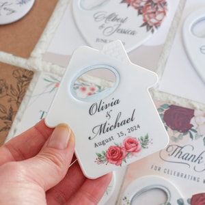 Pode incluir: Abridor de garrafas de cer&acirc;mica branca em forma de casa com um design floral e o texto "Olivia & Michael August 15, 2024".
