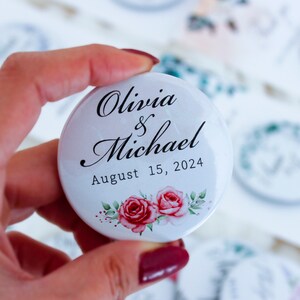 May include: White round button with the text "Olivia & Michael August 15, 2024" in black script font. The button has a floral design with two pink roses.