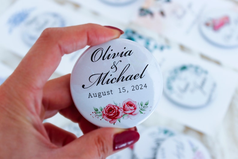 May include: White round button with the text "Olivia & Michael August 15, 2024" in black script font. The button has a floral design with two pink roses.