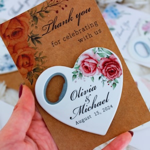 May include: White heart-shaped bottle opener with a floral design and the text "Olivia & Michael August 15, 2024". The bottle opener is on a brown card with the text "Thank you for celebrating with us".