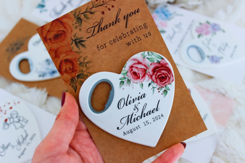 May include: White heart-shaped bottle opener with a floral design and the text "Olivia & Michael August 15, 2024". The bottle opener is on a brown card with the text "Thank you for celebrating with us".
