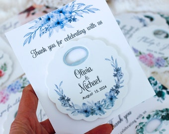May include: White wedding favour with a blue floral design and a bottle opener. The favour says "Olivia & Michael" and "August 15, 2024".