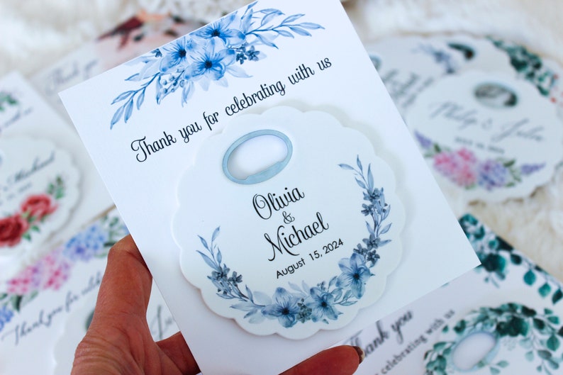 May include: White wedding favor with a blue floral design and a bottle opener. The favor says "Olivia & Michael" and "August 15, 2024".