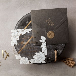 May include: Elegant wedding invitation suite featuring a clear acrylic invitation with white floral design and gold accents. Includes a dark gray envelope with gold initials and a wax seal. Additional details include pencils, rings, and a decorative feather.