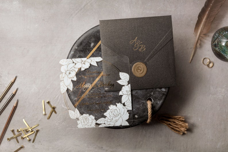 May include: Elegant wedding invitation suite featuring a clear acrylic invitation with white floral design and gold accents. Includes a dark gray envelope with gold initials and a wax seal. Additional details include pencils, rings, and a decorative feather.