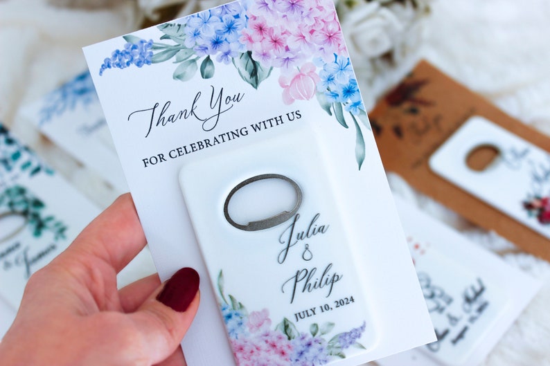 May include: White wedding favor with a floral design and a bottle opener. The favor says "Thank You for celebrating with us" and "Julia & Philip July 10, 2024".