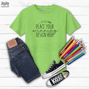 May include: A lime green t-shirt with the text "Place Your Winning Design Here" in black. The t-shirt is laid flat on a white wooden surface with a pair of blue jeans, black sneakers, and a pile of coloured pencils.