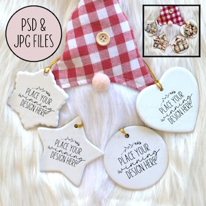 Puede incluir: Adornos de cerámica blanca en varias formas, incluyendo una estrella, un corazón y un círculo, con colgadores de cuerda dorada. Cada adorno tiene el texto "PLACE YOUR WINNING DESIGN HERE". Un gorro de gnomo con un patrón de cuadros rojos y blancos es visible.