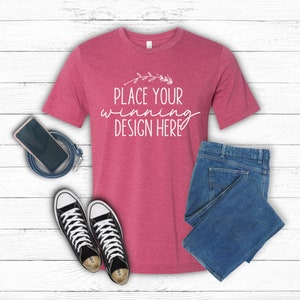 Puede incluir: Una camiseta de color rojo jaspeado con mangas cortas y el texto "PLACE YOUR WINNING DESIGN HERE" en blanco. La camiseta está colocada sobre un fondo de madera blanca con un par de zapatillas negras de caña alta, un par de pantalones vaqueros azules y un teléfono móvil negro con un cable negro.
