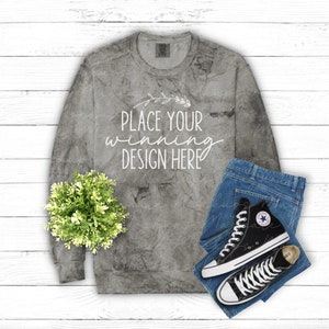 Może przedstawiać: Szara koszula z długim rękawem z wzorem tie-dye i tekstem "Place your winning design here". Koszula leży płasko na białym drewnianym tle z niebieskimi dżinsami i czarnymi wysokimi butami.