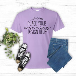 May include: A lilac-colored t-shirt with the text "PLACE YOUR WINNING DESIGN HERE" in black script. The shirt is paired with blue jeans and black canvas sneakers with white laces. A small potted plant with green and white leaves is also present.